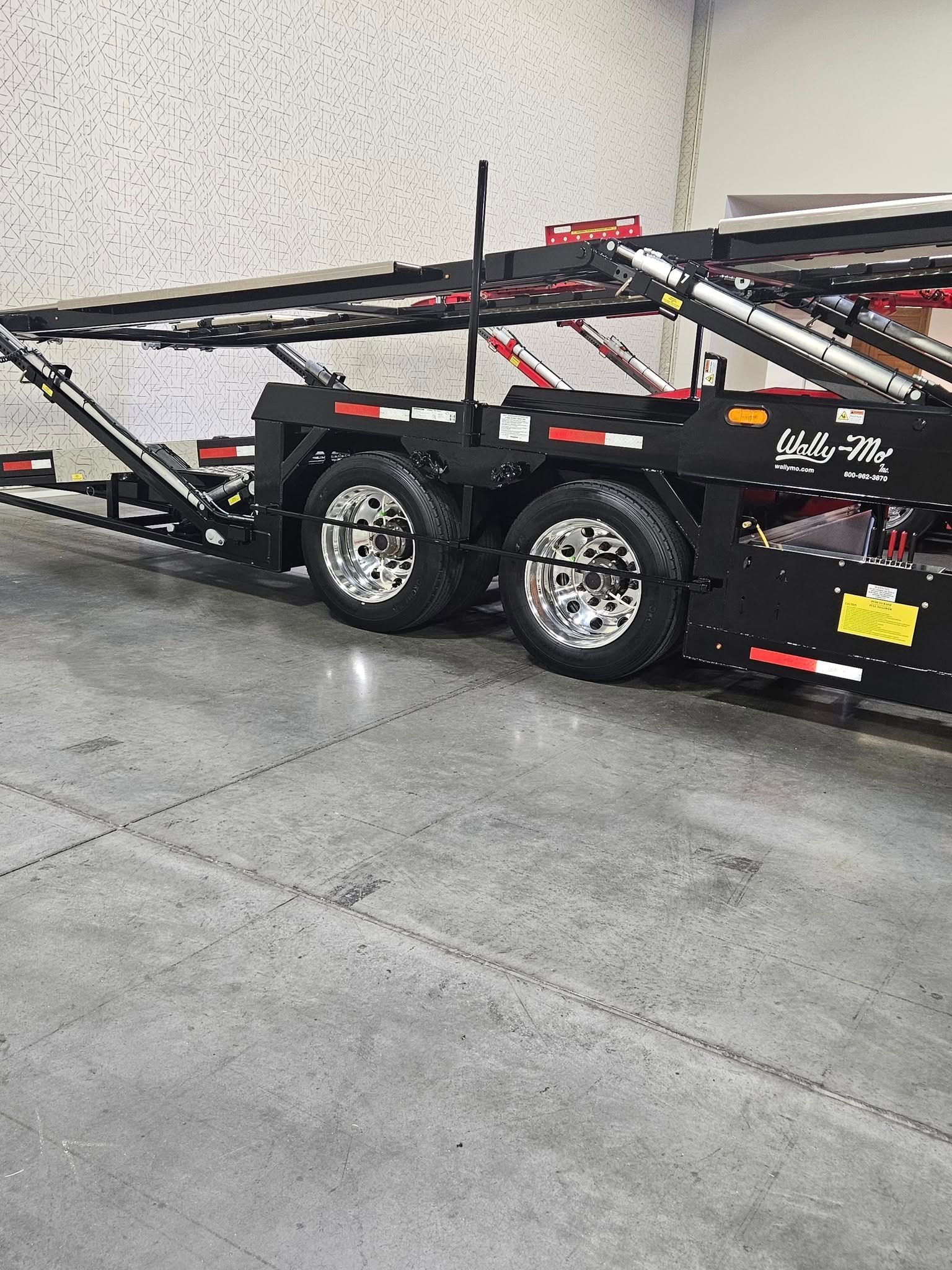 A black car hauler trailer with chrome wheels parked on a gray concrete floor inside a large warehouse.