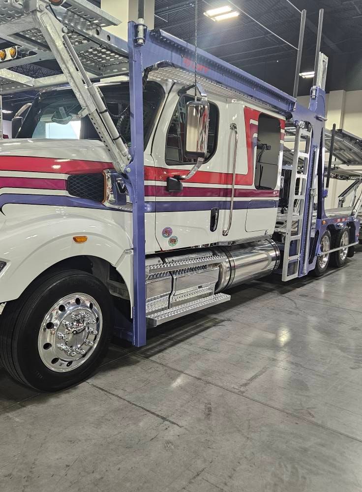 A white, red, and purple car hauler truck parked inside a warehouse with metal loading ramps over the cab.