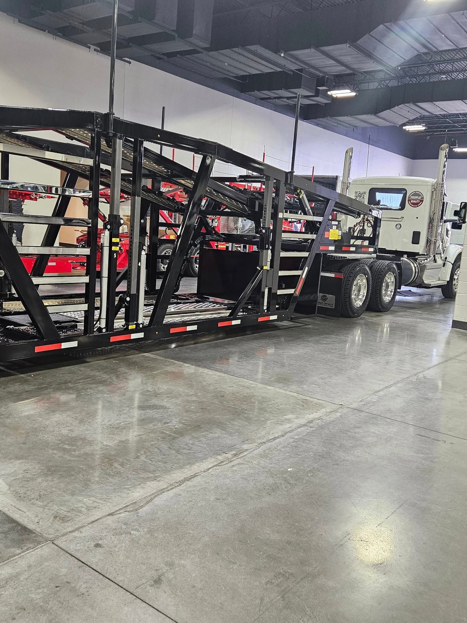 A white semi-truck pulling a large, black metal car hauler trailer inside a warehouse with a polished concrete floor.