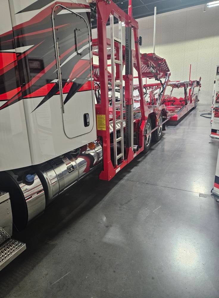 A white semi-truck with red and black graphic designs connected to an empty red car carrier trailer inside a garage.