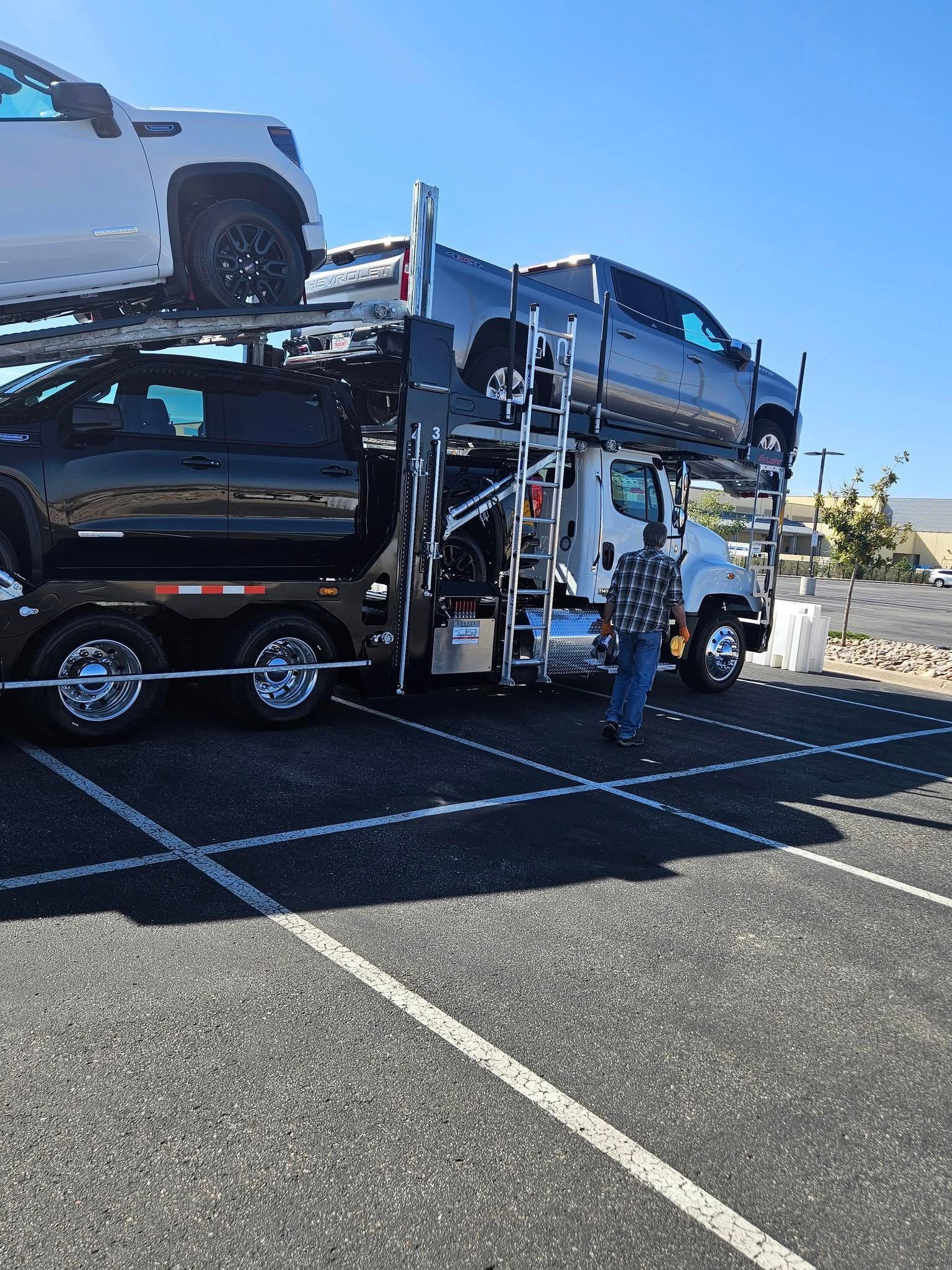 A truck driver stands next to a car hauler loaded with several new pickup trucks in a paved parking lot under a blue sky.