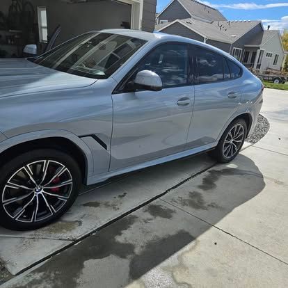 A light grey BMW X6 SUV parked on a residential concrete driveway on a sunny day.