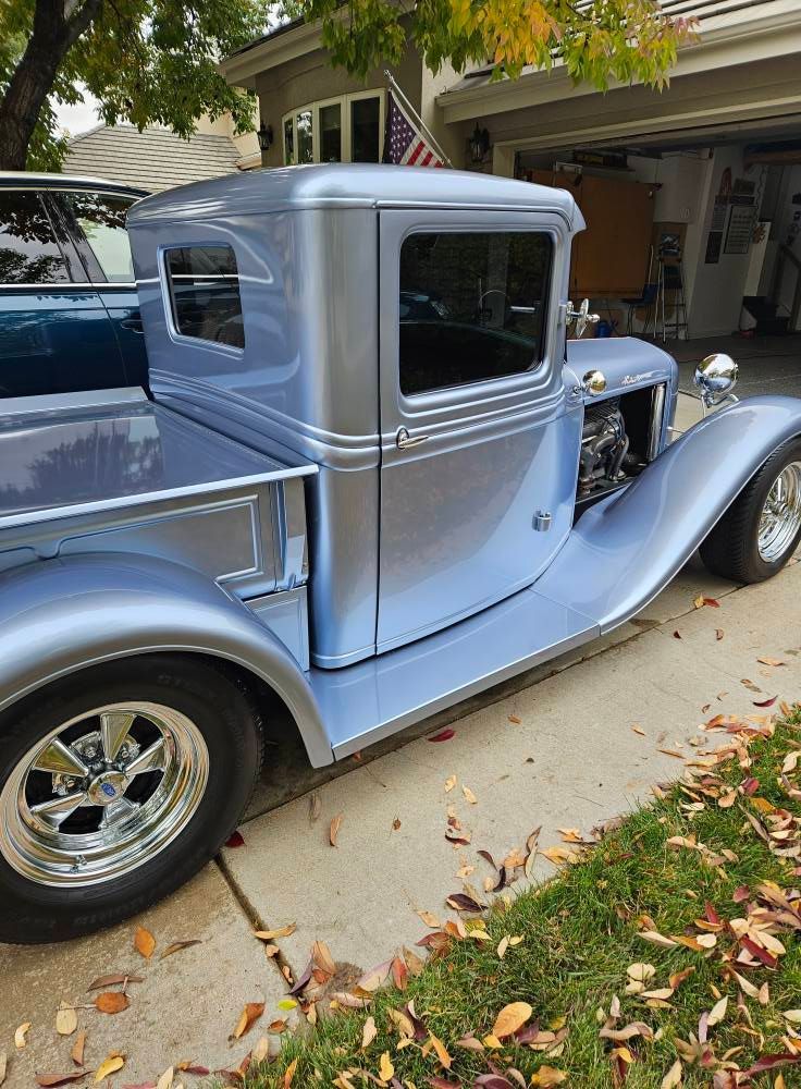 A side view of a light blue custom hot rod truck parked in a driveway next to a house with an American flag.
