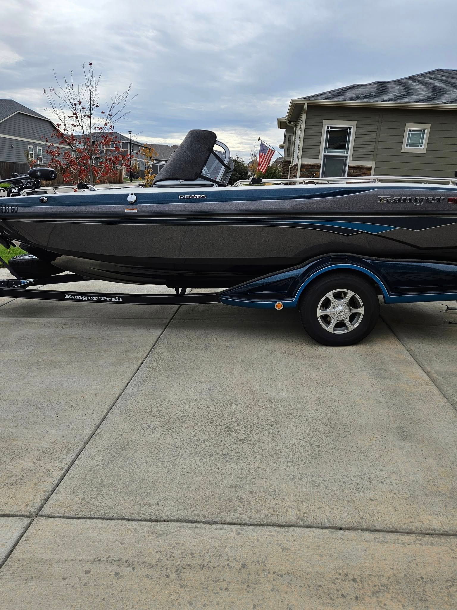 A dark-colored bass boat on a blue trailer parked on a concrete driveway in front of a suburban house.