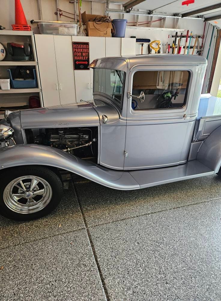 A classic silver vintage hot rod truck parked inside a cluttered residential garage.