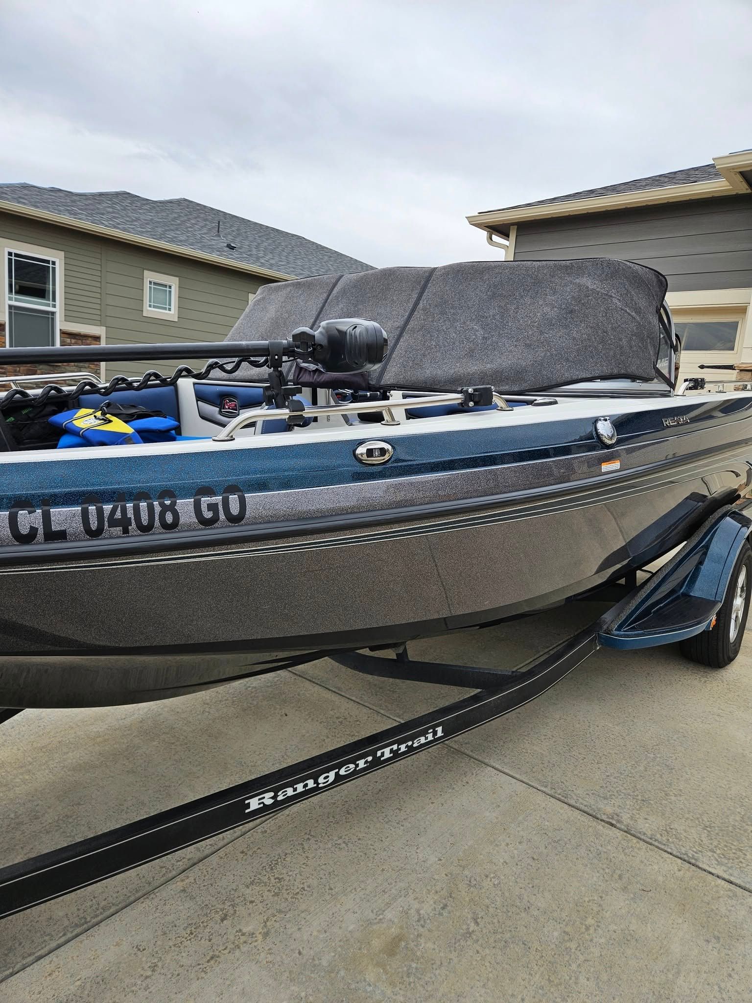 A dark gray bass boat on a trailer parked on a concrete driveway in front of a residential house.