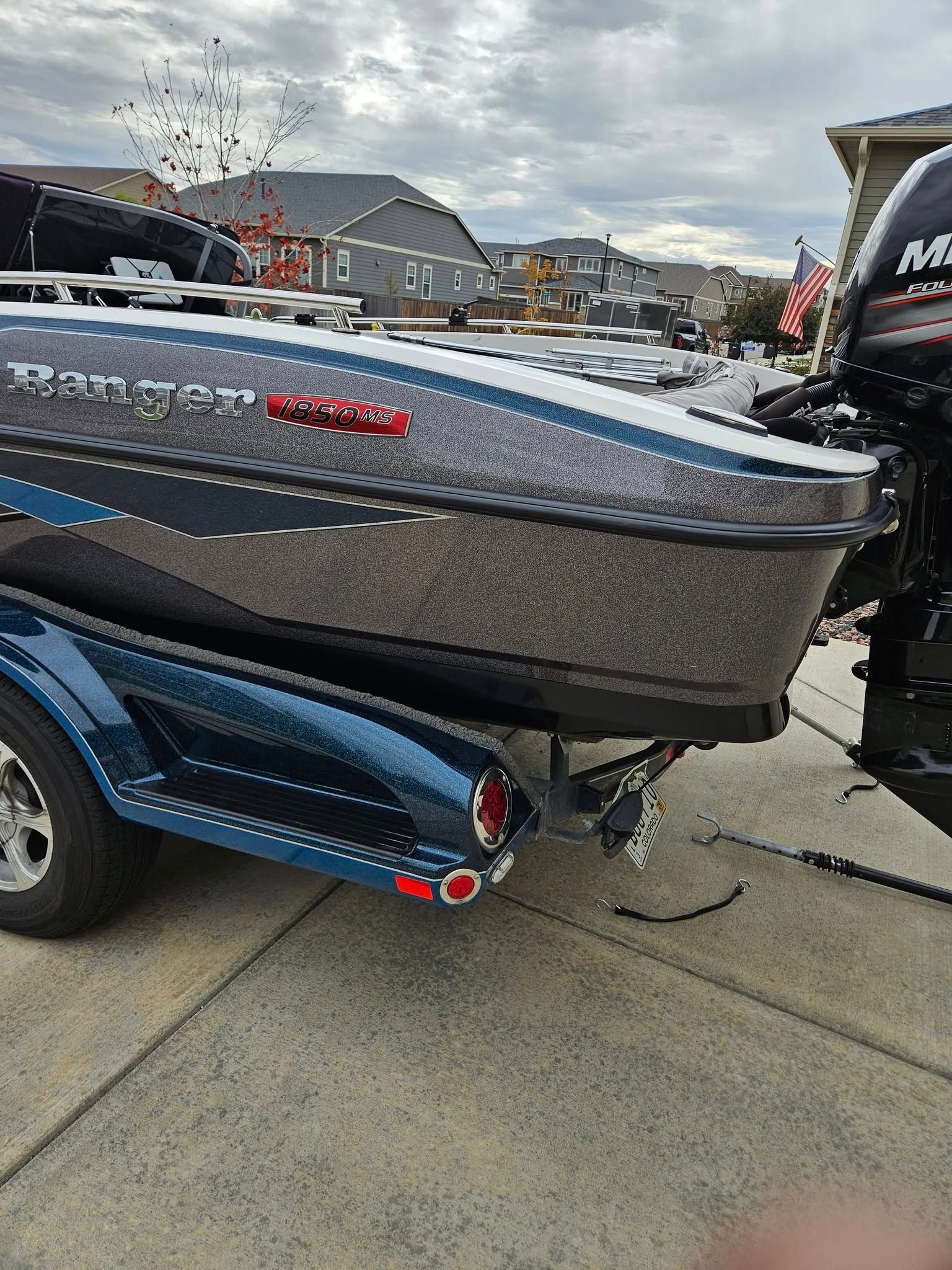 A grey and blue bass boat on a trailer parked on a concrete driveway in a suburban neighborhood.