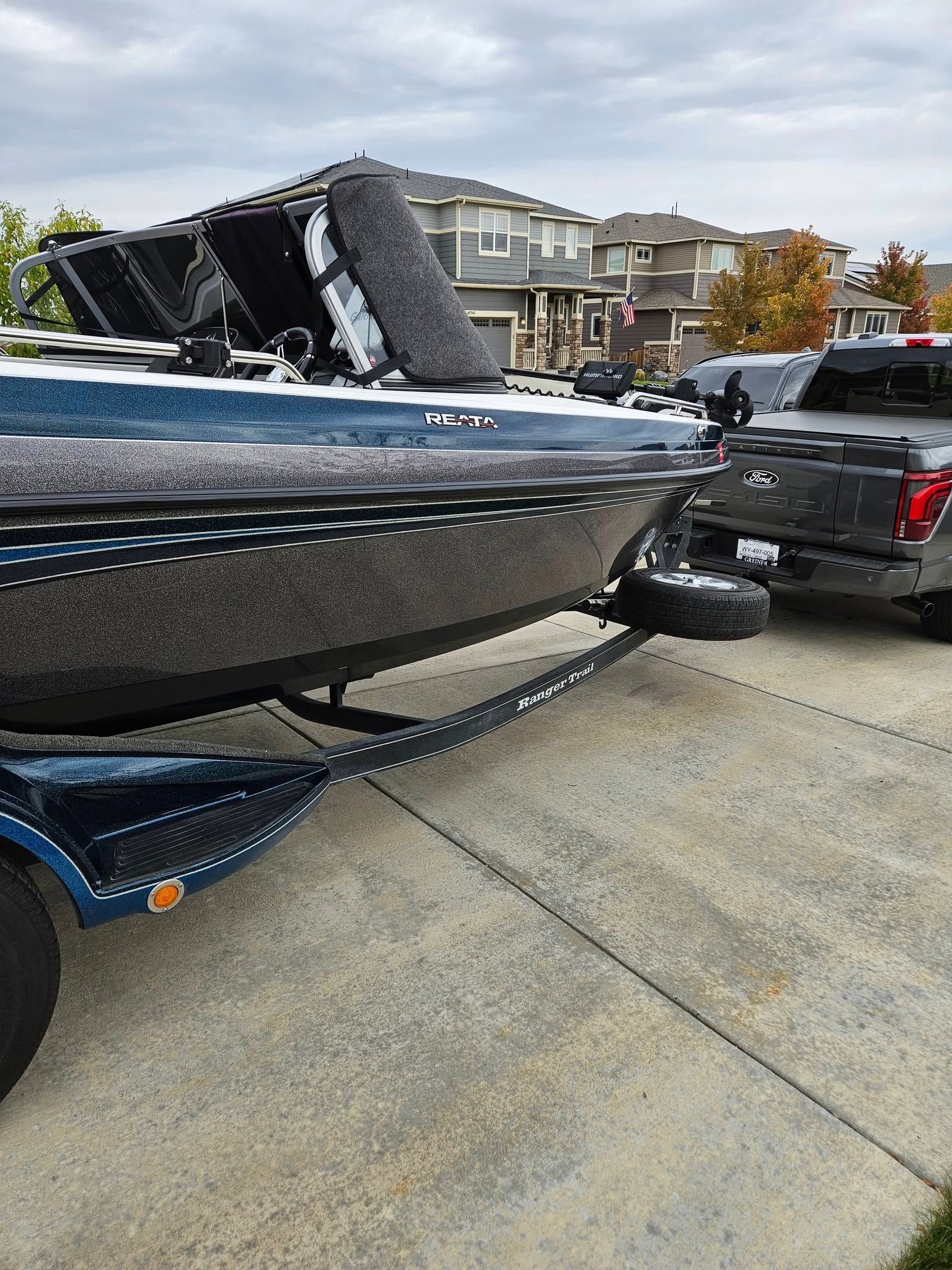 A dark blue bass boat on a trailer parked in a residential driveway next to a grey pickup truck.