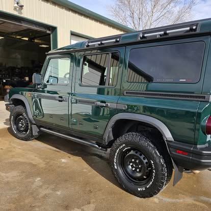 A dark green four-door Jeep Wrangler parked outside a shop, viewed from the rear passenger side.