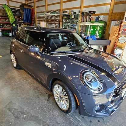 A dark grey Mini Cooper parked inside a workshop with wooden walls, metal shelves, and a green banner.