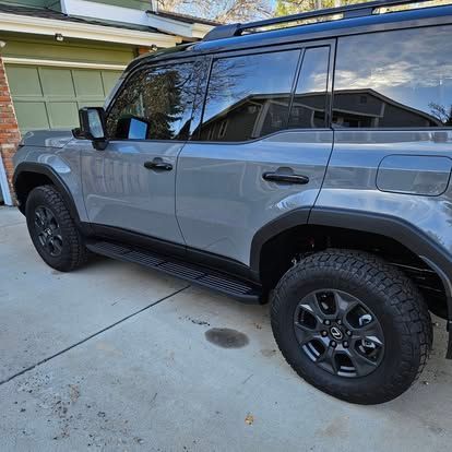 A gray Lexus GX 550 SUV parked in a driveway, viewed from the side, showing its off-road wheels and black trim.