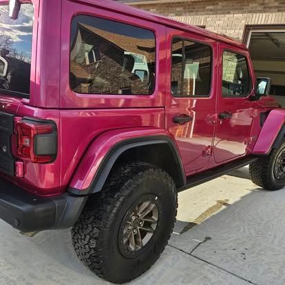 A bright magenta four-door Jeep Wrangler parked on a concrete driveway in front of a brick house.