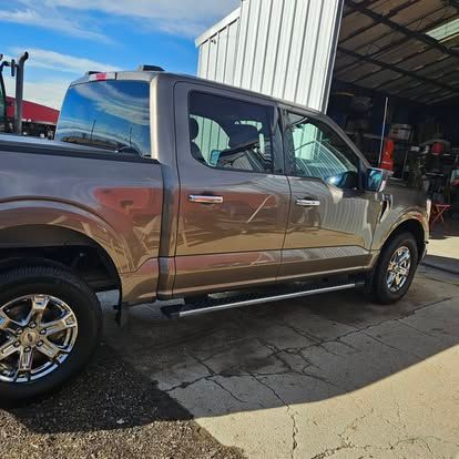 A brown four-door Ford pickup truck parked on a concrete lot next to a metal building under a blue sky.