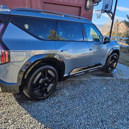 A side view of a gray, modern SUV with black wheels parked on a gravel driveway near a basketball hoop and a red building.