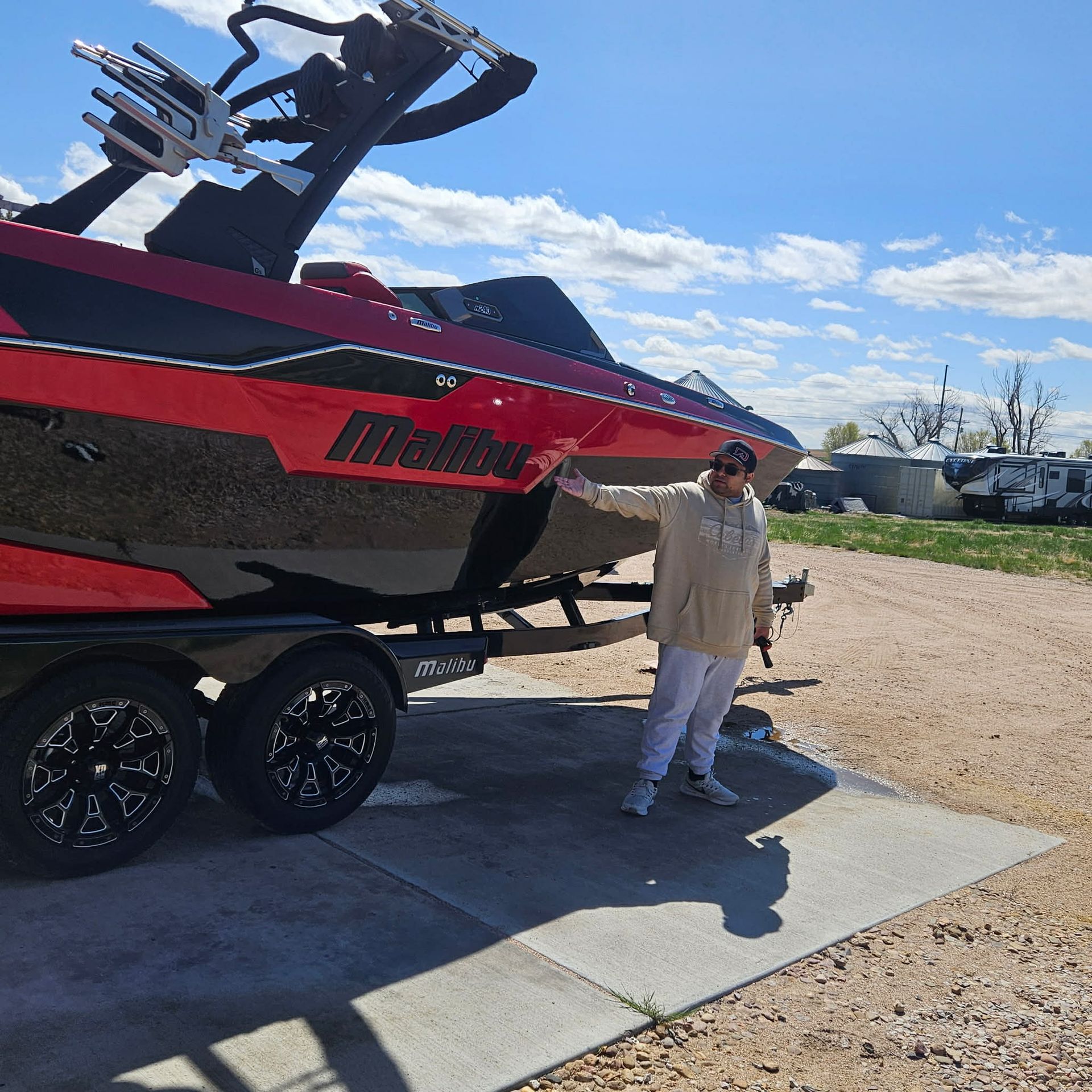 A person wearing a beige hoodie gestures toward a red and black Malibu motorboat parked on a trailer in a gravel lot.