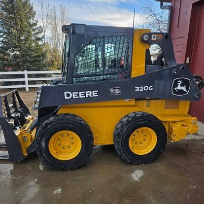 A yellow and black John Deere 320G skid steer loader parked on a concrete surface next to a red building.