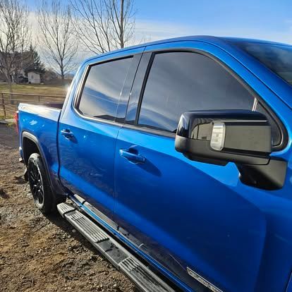 A close-up, angled view of a bright blue pickup truck parked on dirt, showing the tinted windows and black side mirror.