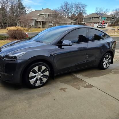 A dark gray Tesla Model Y parked on a concrete driveway in a suburban neighborhood.