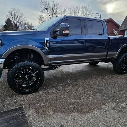 A dark blue Ford F-Series crew cab pickup truck with two-tone tan trim and large black custom wheels parked on gravel.