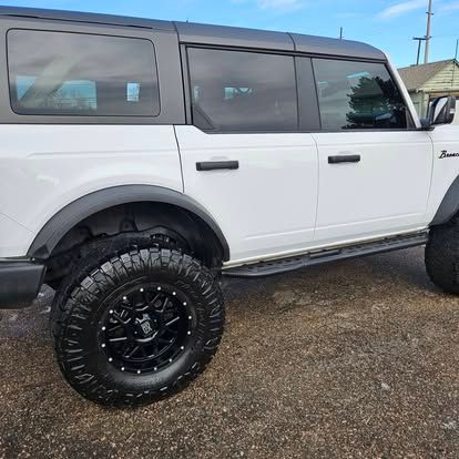 A side view of a white four-door Ford Bronco with black wheels and a black hardtop parked on gravel.