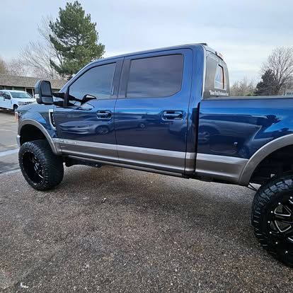 A lifted blue Ford F-Series pickup truck with grey lower trim and black custom wheels parked on an asphalt lot.
