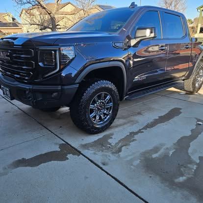 A dark gray GMC Sierra AT4 crew cab pickup truck parked on a concrete driveway in a suburban neighborhood.