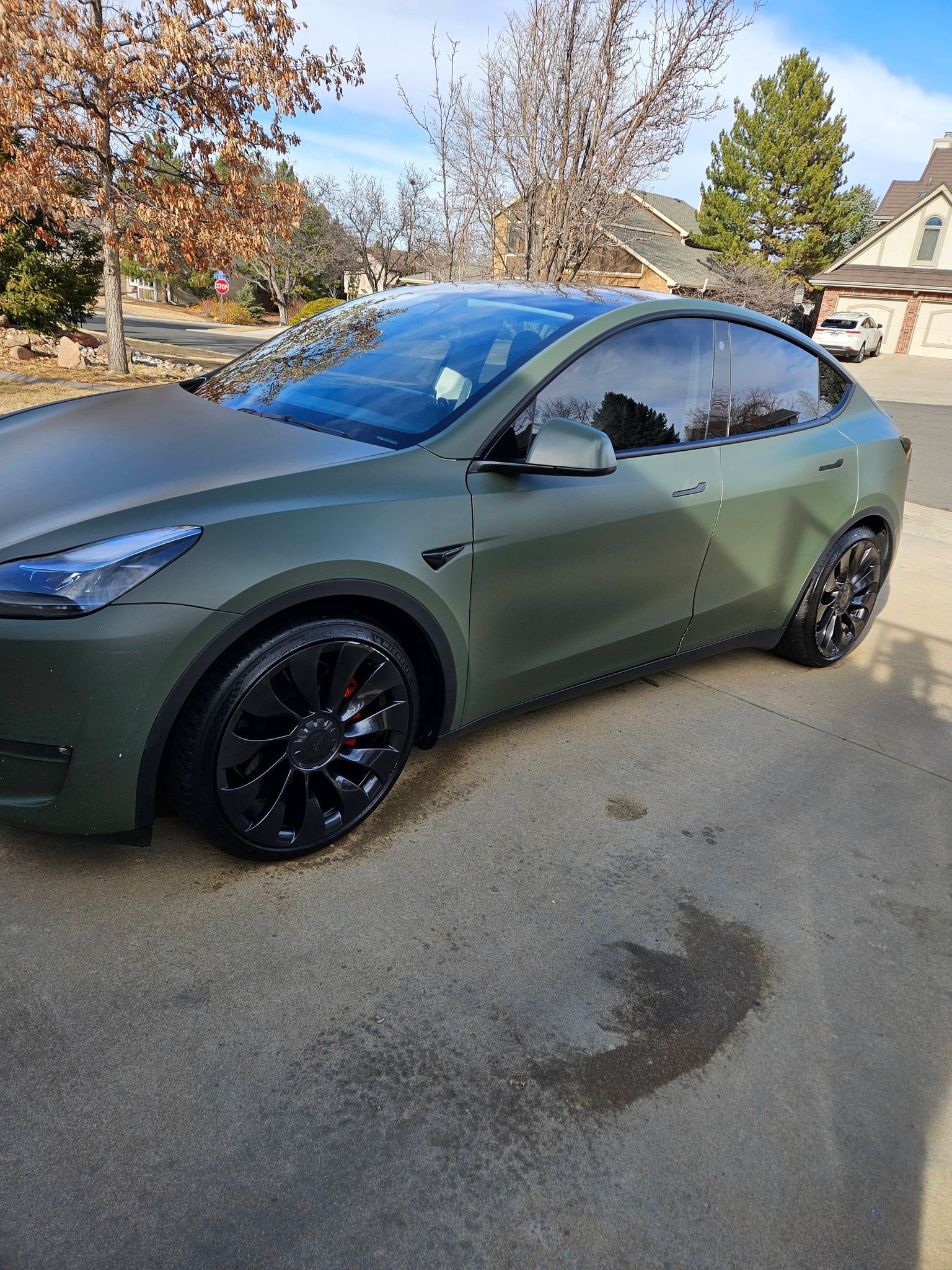 A matte olive-green Tesla Model Y parked on a concrete driveway in a suburban neighborhood.