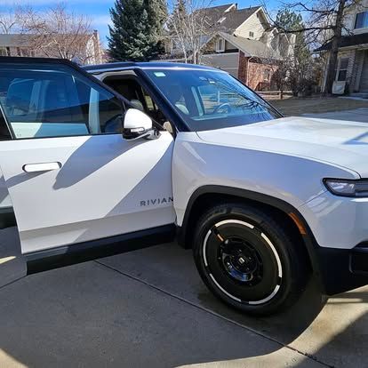 A white Rivian SUV with its driver-side door open, parked on a suburban concrete driveway under a clear blue sky.