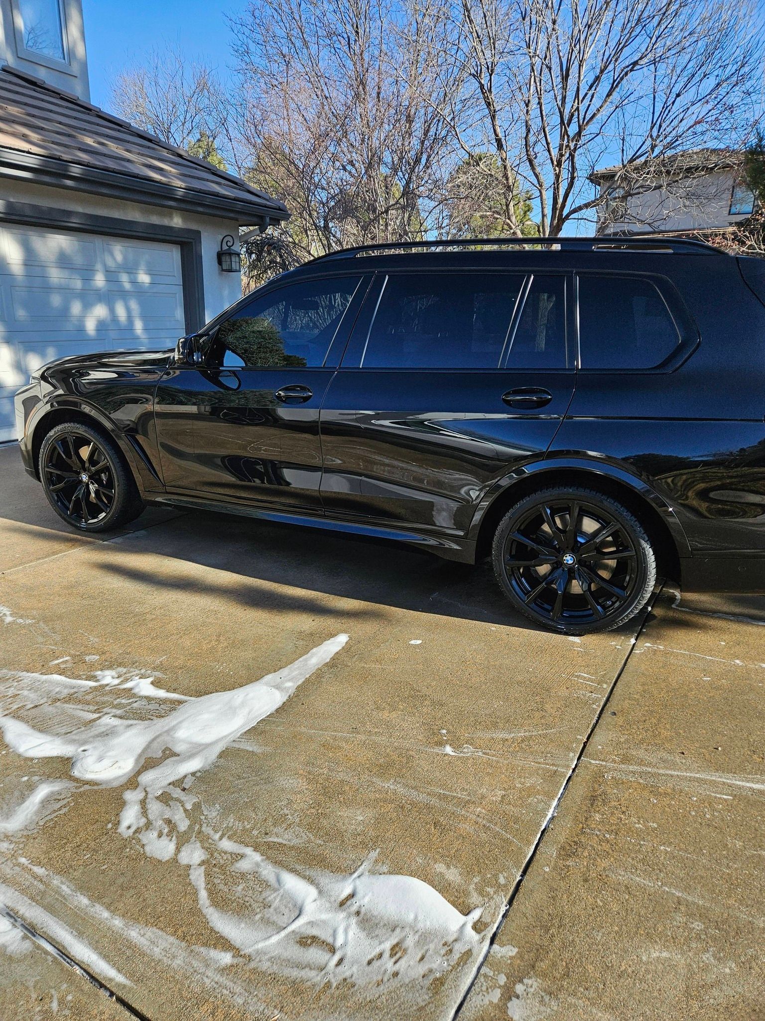 A shiny, black SUV parked in a residential driveway with soapy water foam on the concrete.