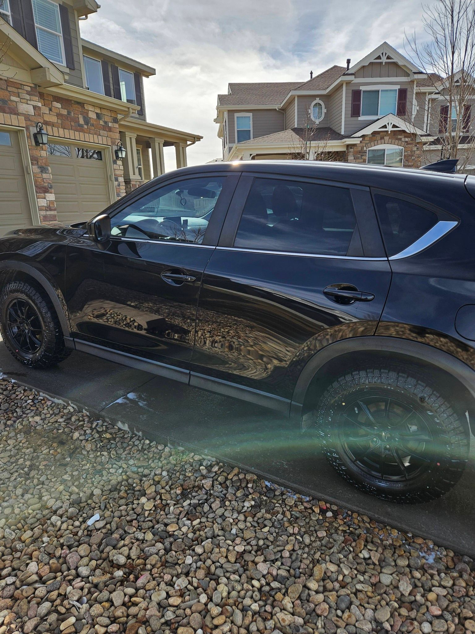 A black SUV parked on a pebble driveway in front of suburban houses on a partly cloudy day.