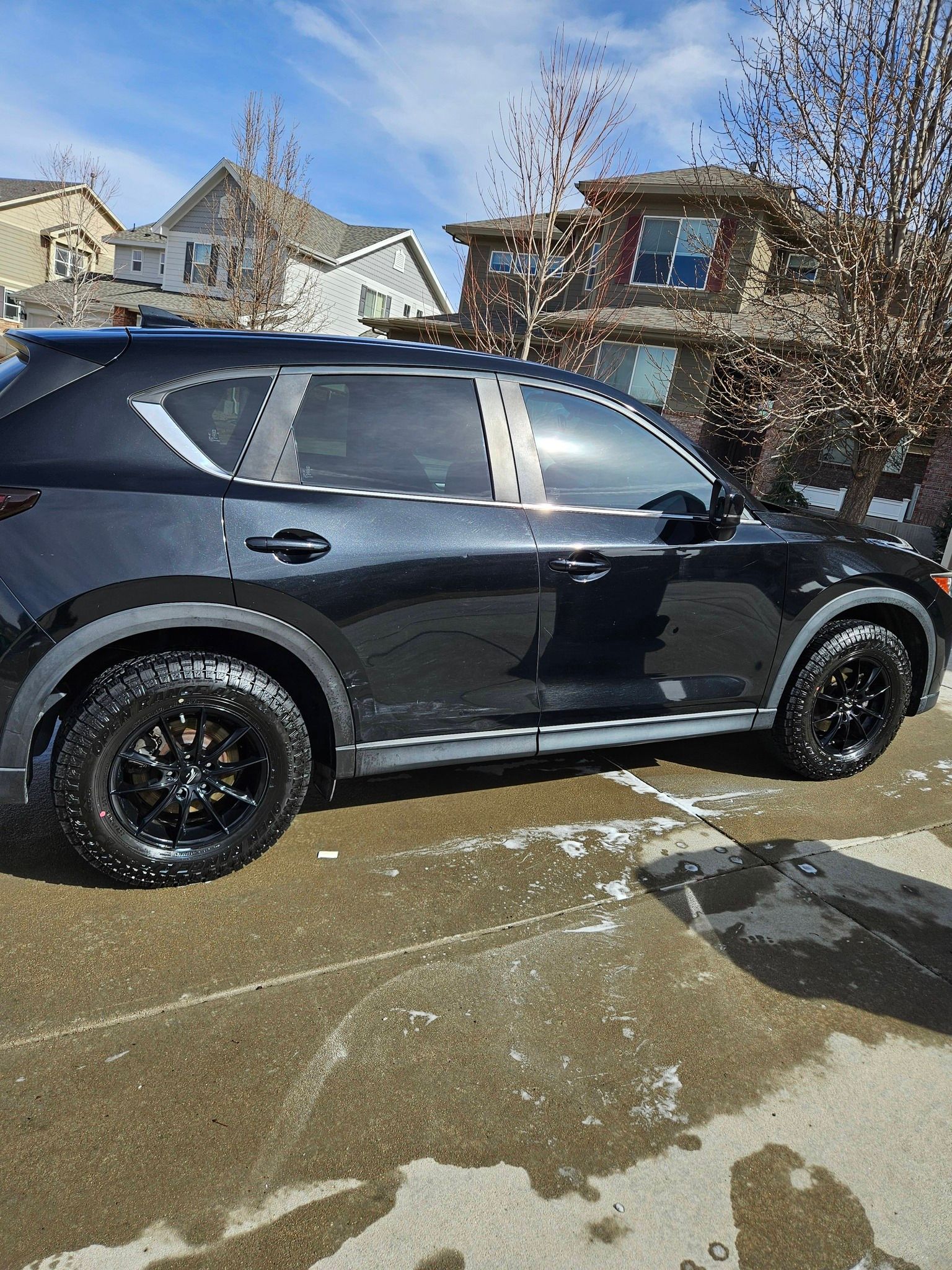 A black SUV with off-road tires parked on a driveway near a suburban house on a sunny day.