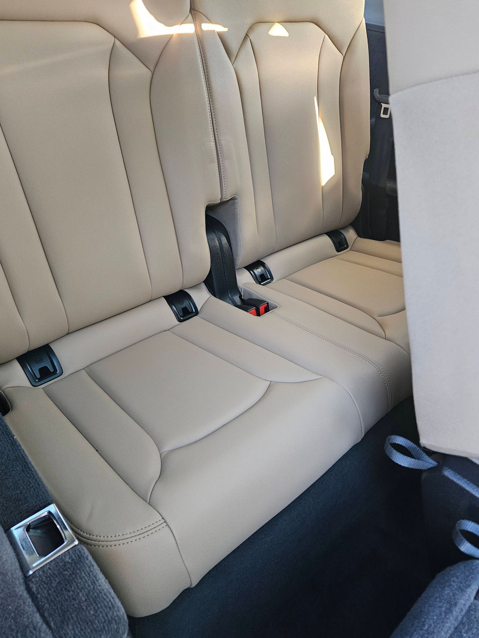 Tan-colored car rear seats with black seat belt buckles and dark gray floor matting.