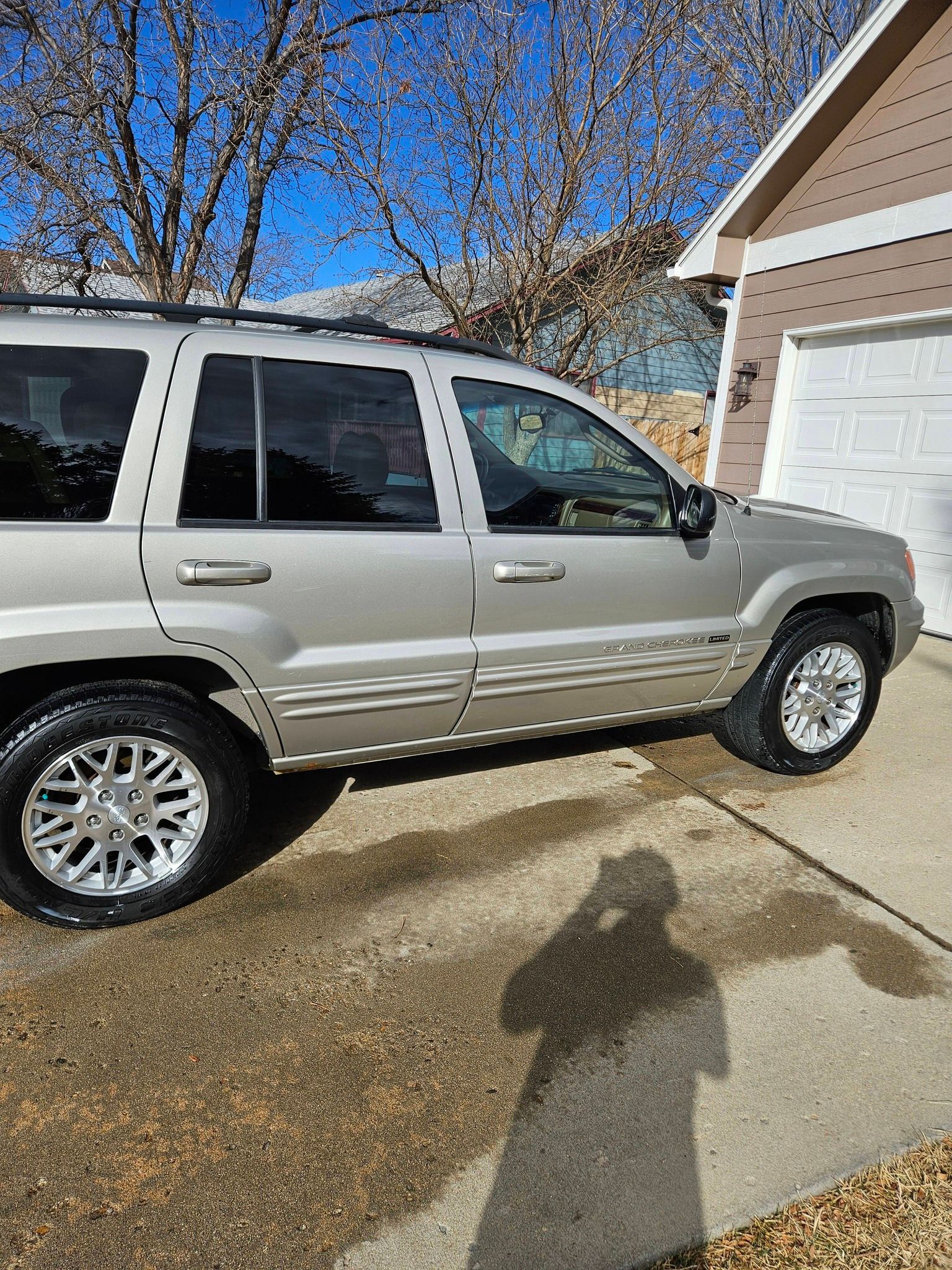 A beige Jeep Grand Cherokee SUV parked on a concrete driveway in front of a house on a sunny day.