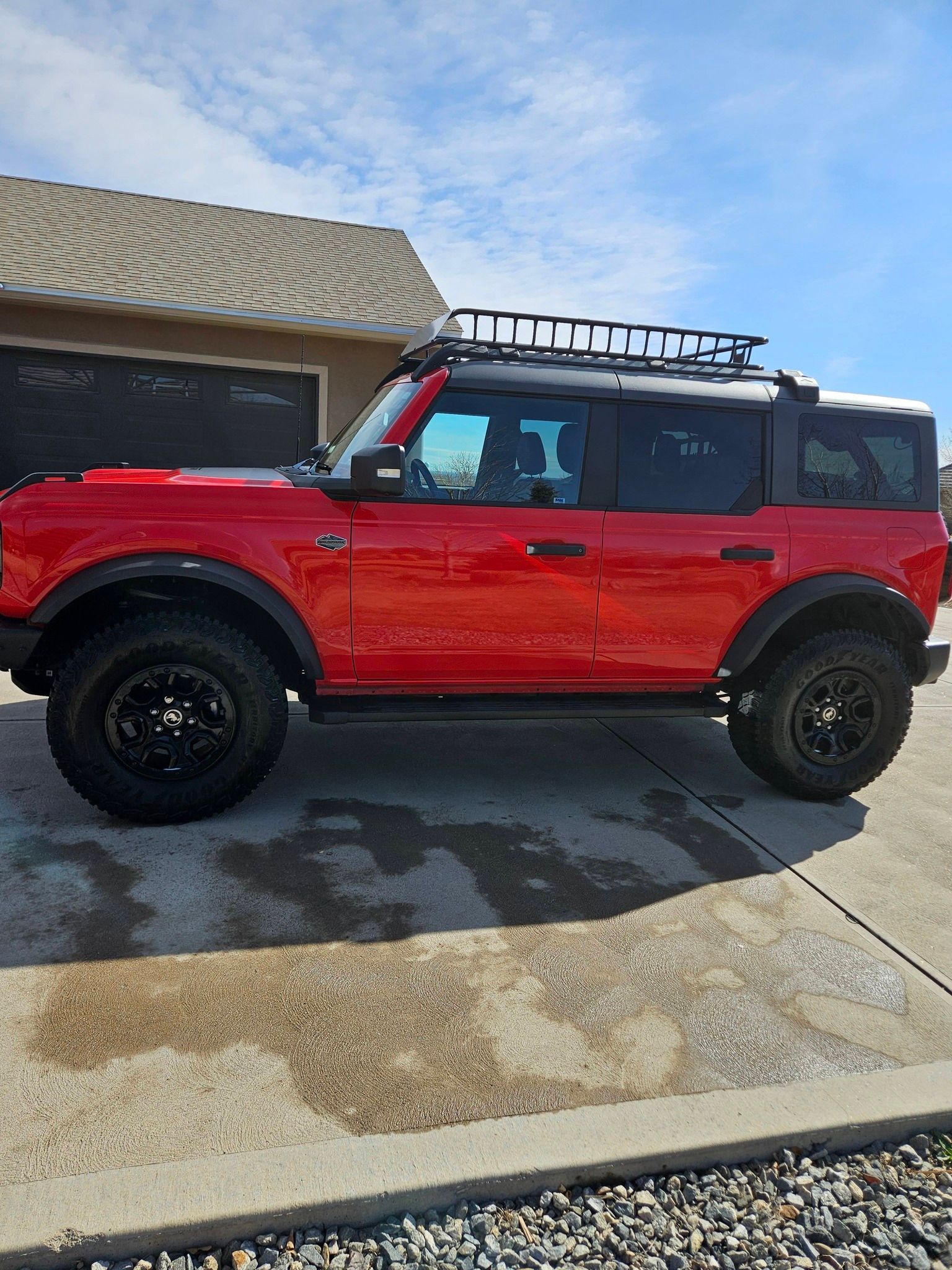 A red Ford Bronco with a roof rack and black wheels parked on a concrete driveway in front of a house.