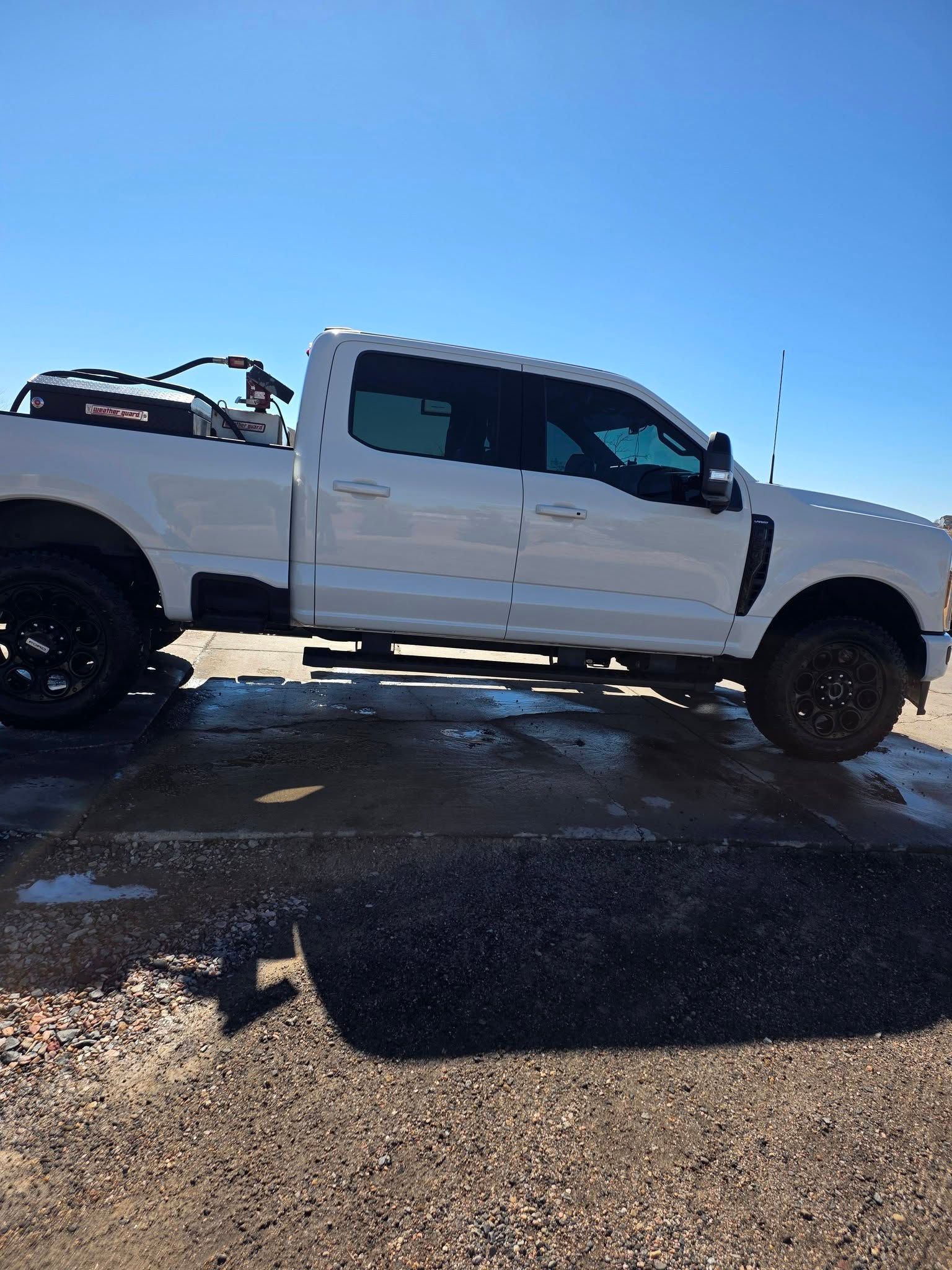 A white Ford F-Series pickup truck with black wheels and an equipment tank in the bed parked on a gravel lot.