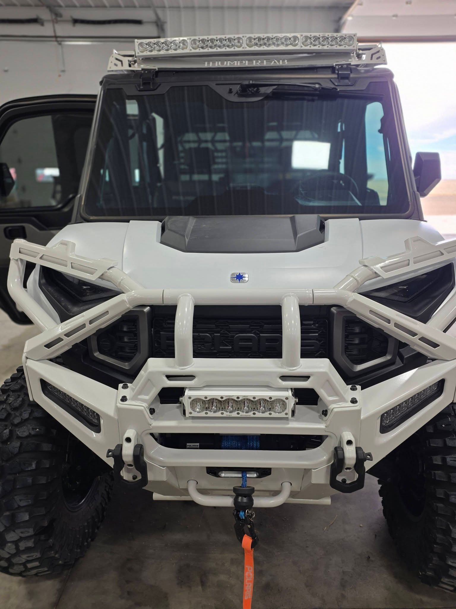 A white off-road utility vehicle with a heavy-duty custom bumper, LED light bars, and winch in a garage.