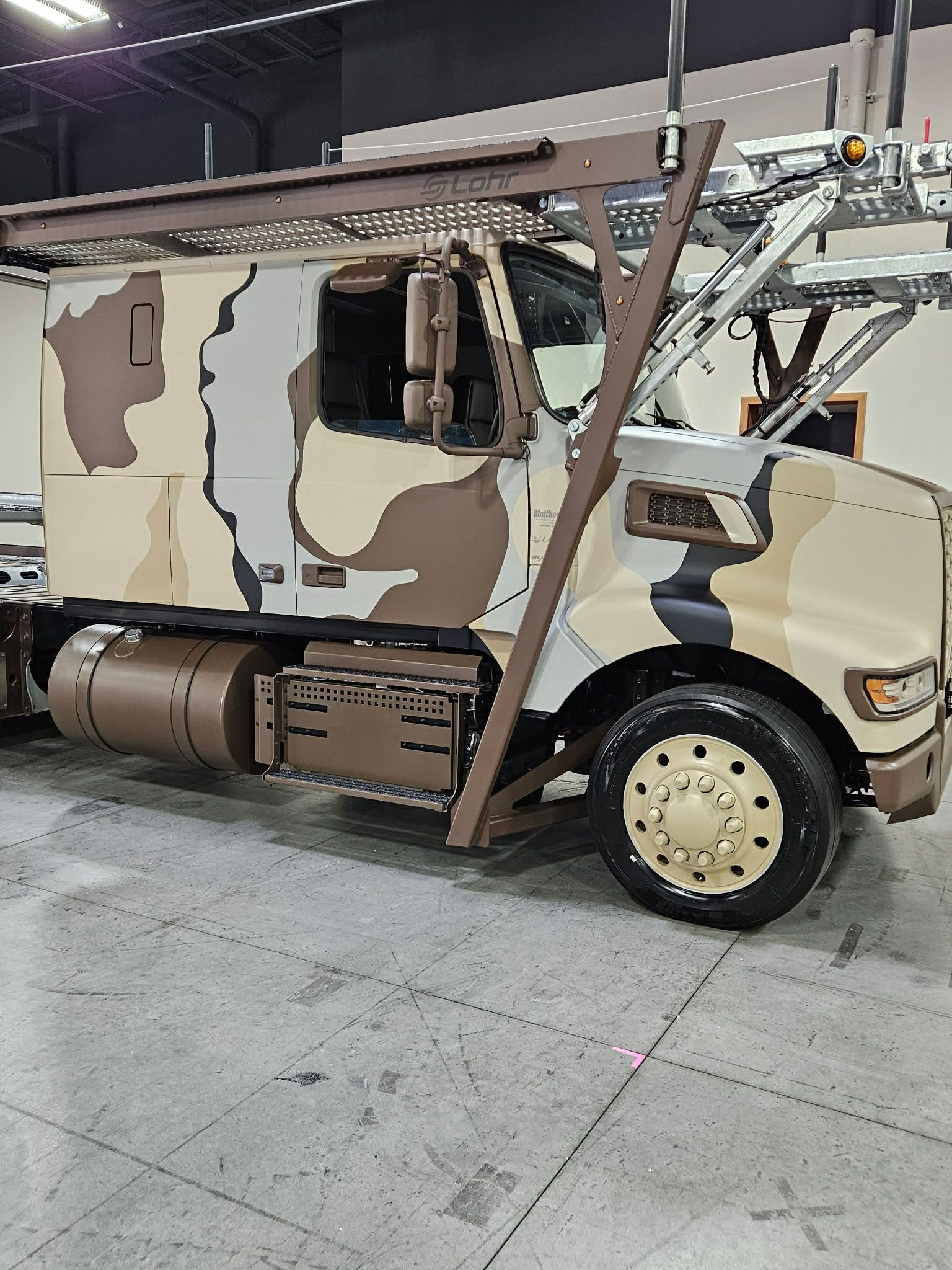 A semi-truck with a tan, brown, and grey camouflage paint job parked indoors with a car hauler rack attached.