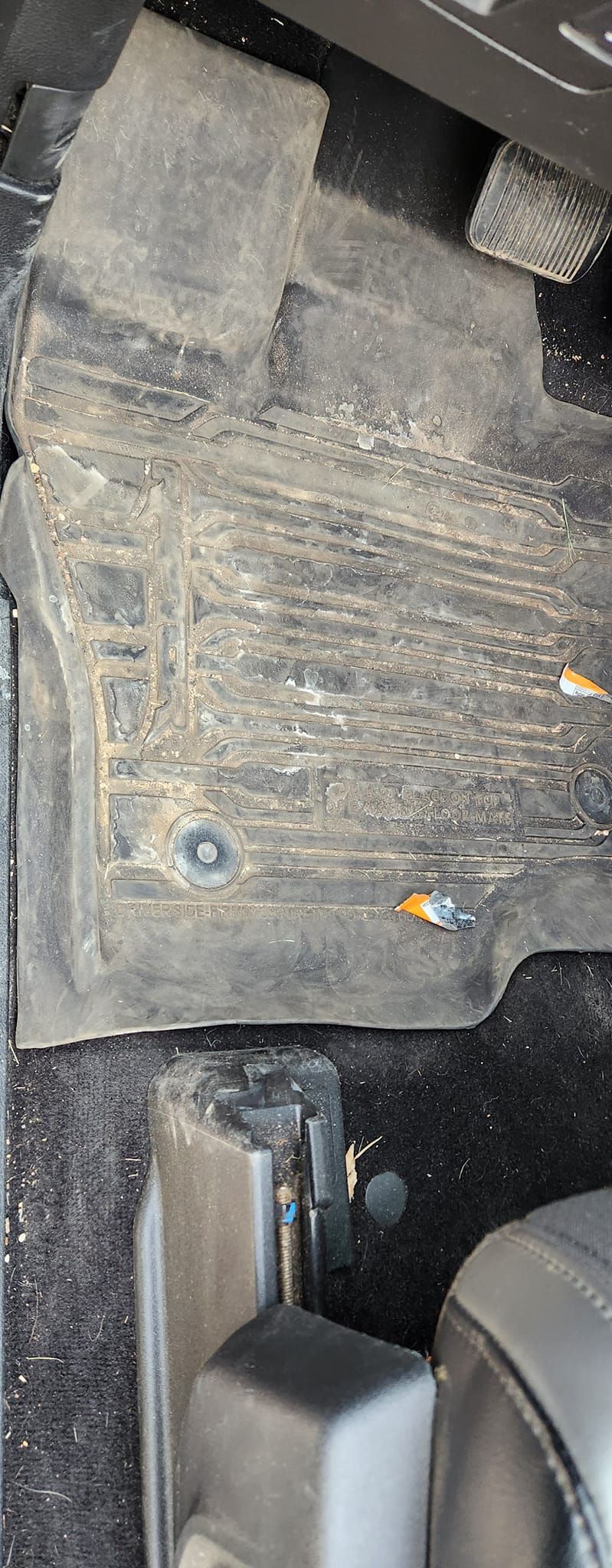A dirty, light-colored car floor mat on a black carpet, showing wear, debris, and two metal anchor grommets.