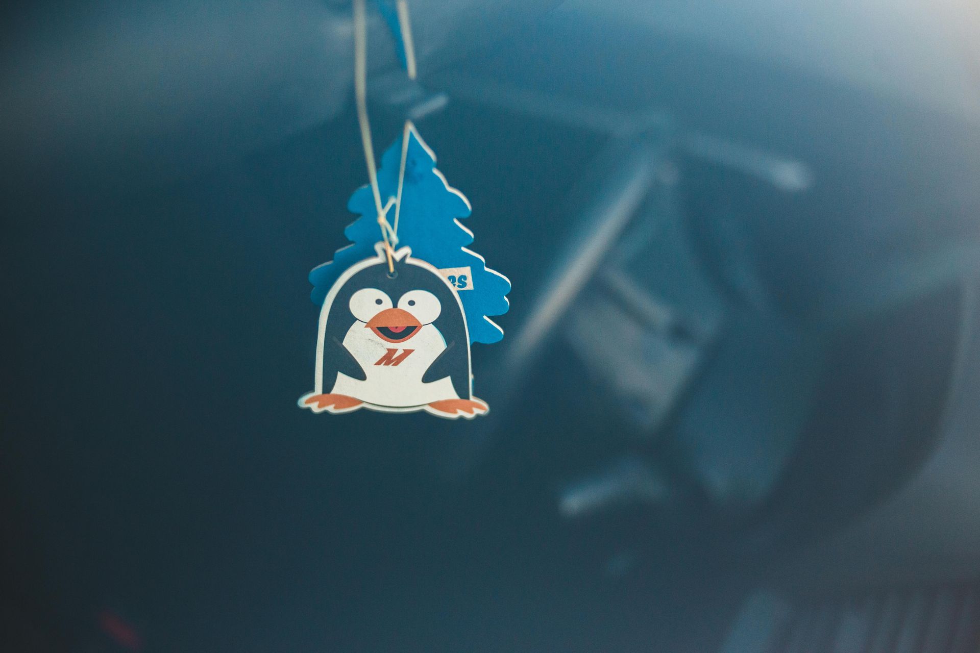 A car air freshener shaped like a blue pine tree with a cartoon penguin graphic attached, hanging inside a vehicle.