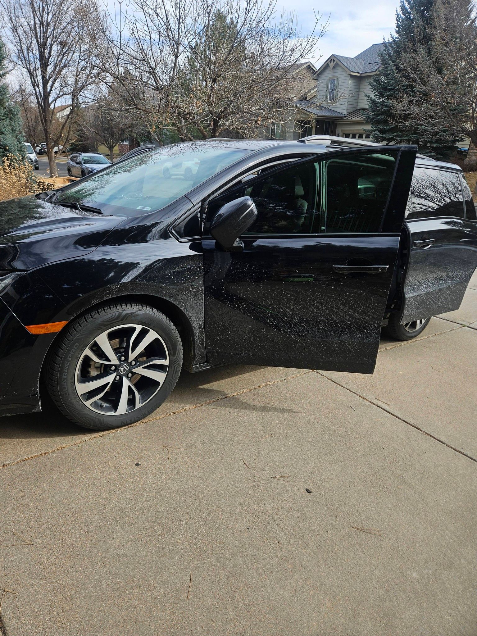 A black minivan parked on a residential driveway with its front passenger door open, covered in light snow or frost.