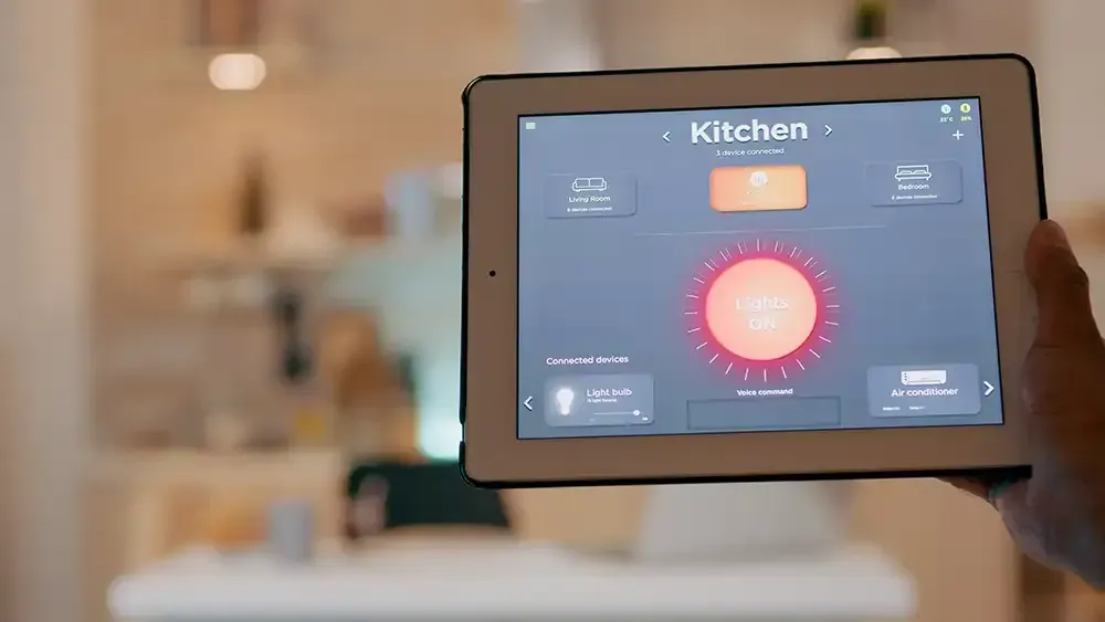 A pic of a hand holding a tablet inside a home. The tablet screen shows a light control system that is controlling the kitchen light