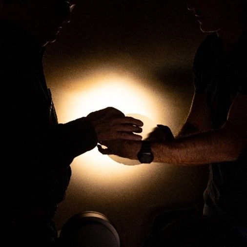 Two individuals in a dark room passing a light source, symbolizing collaboration or sharing knowledge