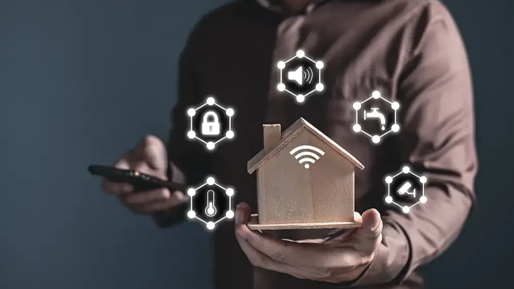 A man holding a phone in one hand and a mock-up of a house in another. Indicating that the phone can control multiple features of the house