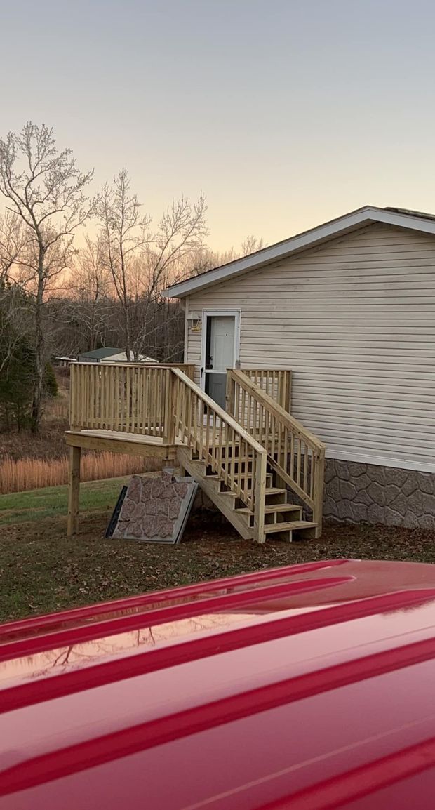 A wooden deck with stairs attached to the side of a light-colored mobile home, seen from across a grassy yard at sunset.