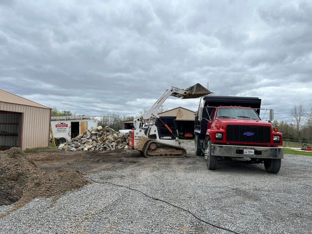 A white skid steer loader lifts a bucket of debris toward a red dump truck parked on a gravel lot next to outbuildings.