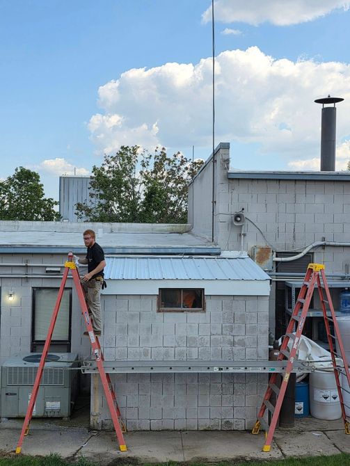A person in work clothes stands on a tall orange ladder against a cinder block building with a metal awning.