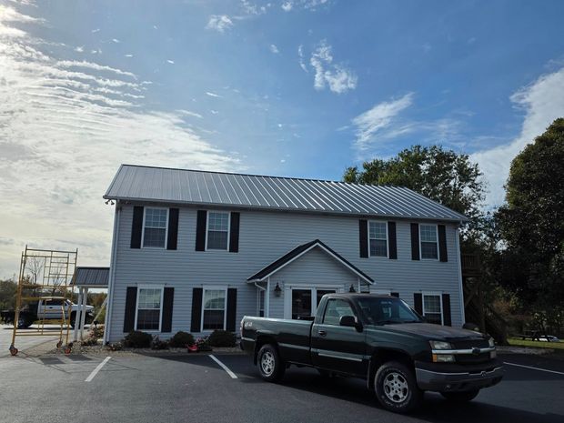 A dark green pickup truck parked in front of a two-story gray house with a newly installed silver metal roof.