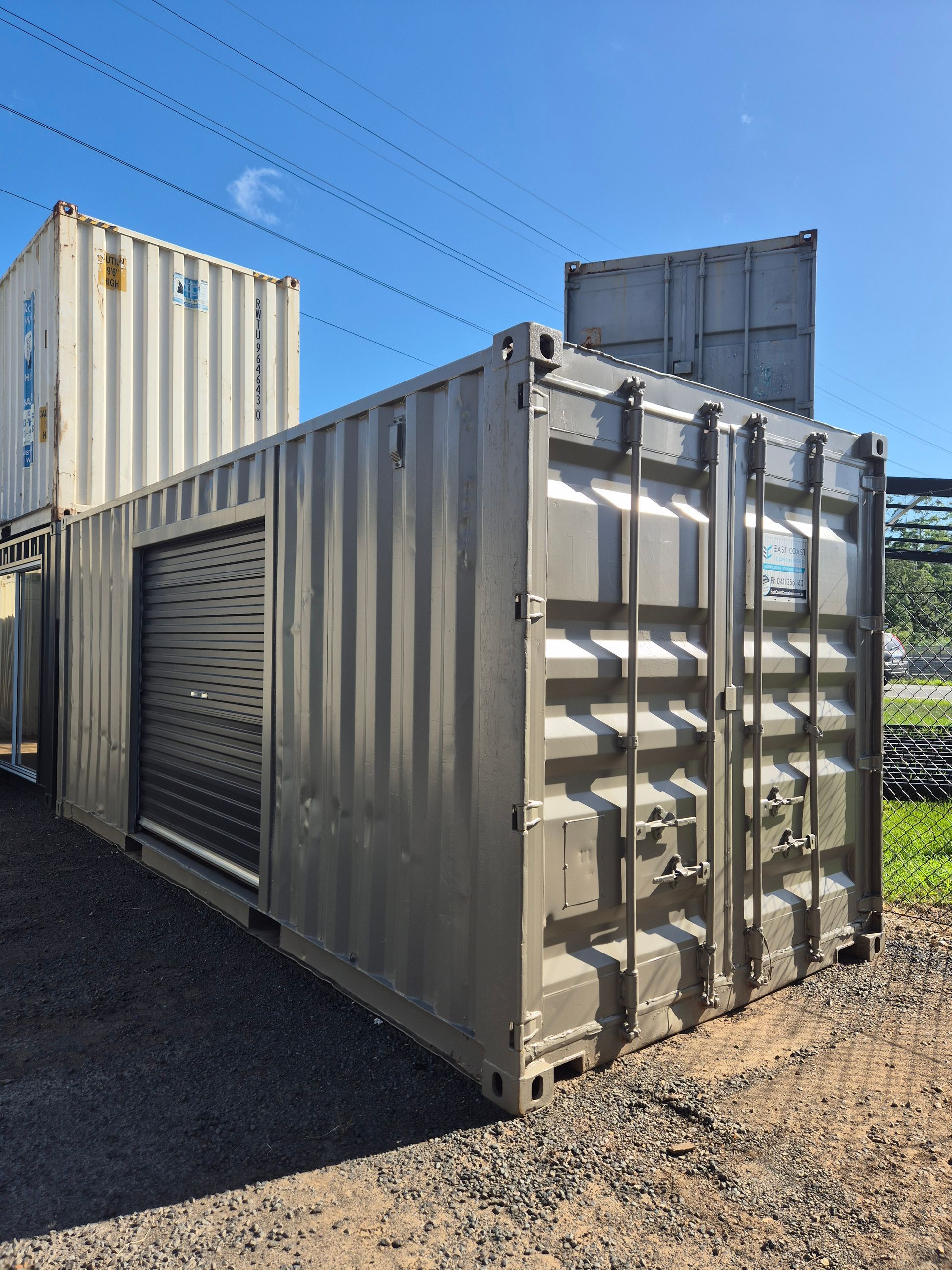 Grey shipping container with roller door beside stacked containers under a clear blue sky.