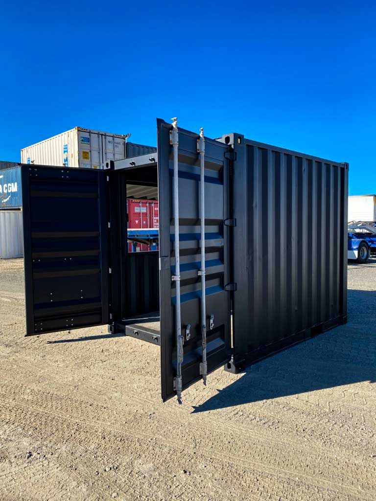 A black shipping container with its doors open is sitting on top of a dirt field.