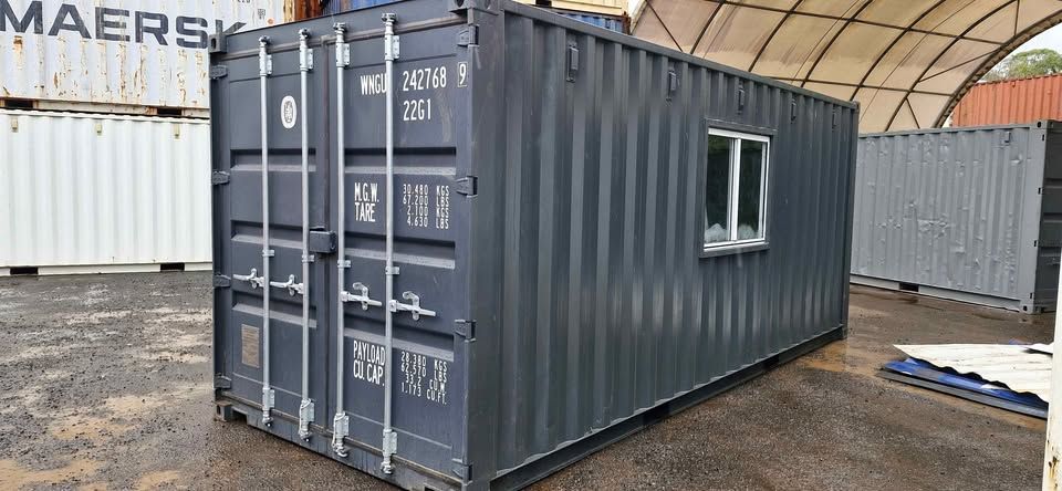 Dark grey shipping container modified with a window, placed in an industrial storage area.